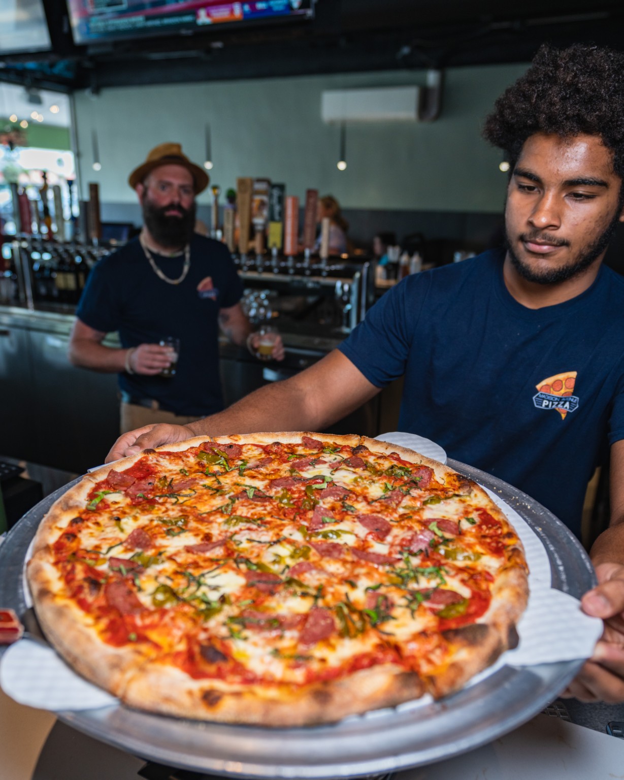 A Madison Avenue Pizza employee presenting a freshly baked pizza, with a colleague behind the bar